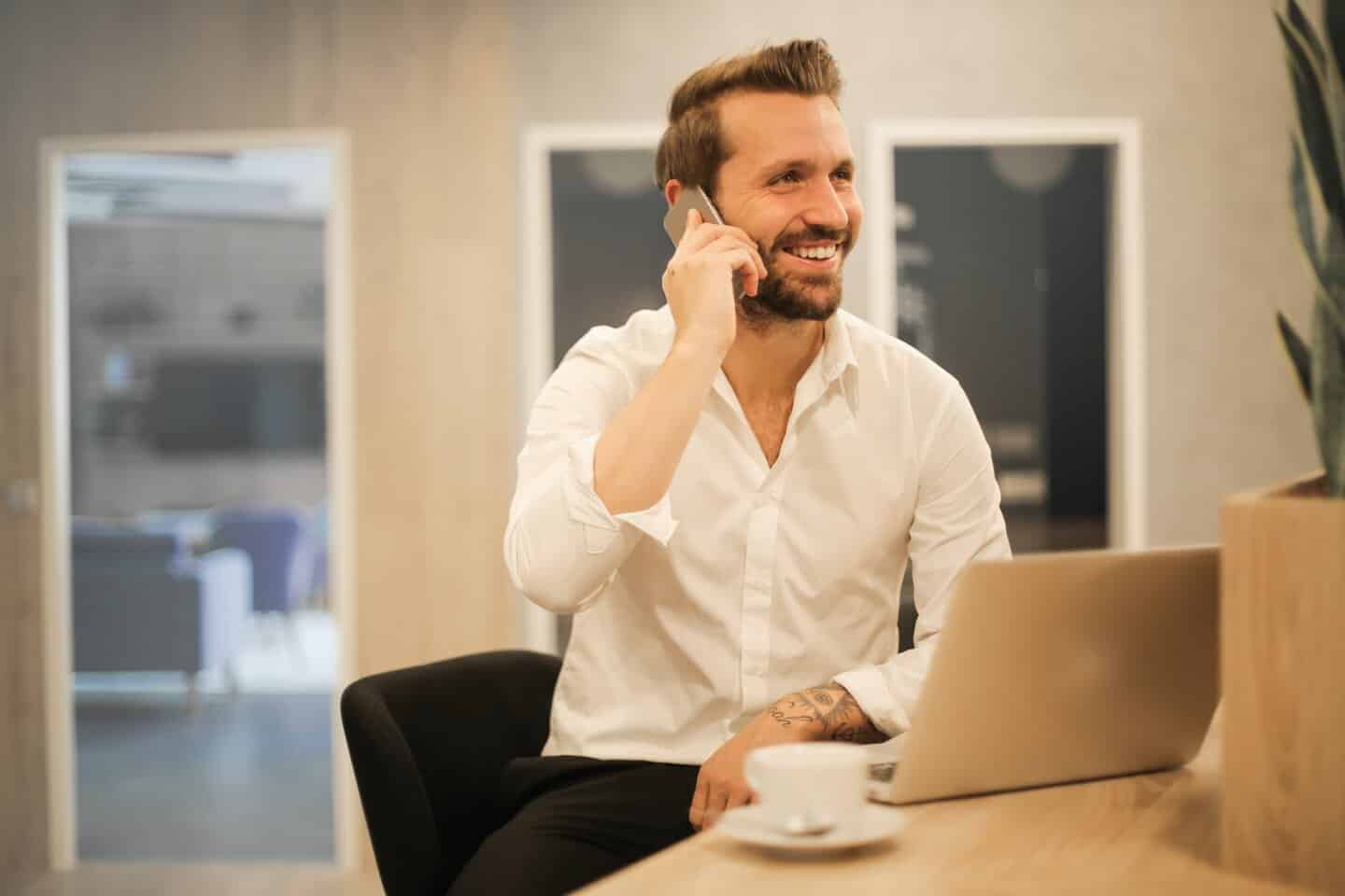 A man in the office in front of laptop, he is approximately 30 years old. He has a trimmed beard and holding a mobile phone to his ear. He is a DisabilityPlus therapist using his phone and laptop to take sight loss counselling sessions.