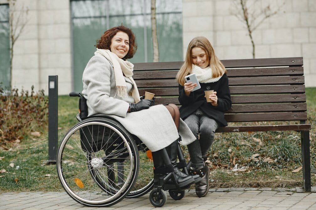 A lady sat in her wheelchair outside. She is sat next to her freind who is looking at the DisabilityPlus website, she is deciding if cerebral palsy counselling is good for her freind.