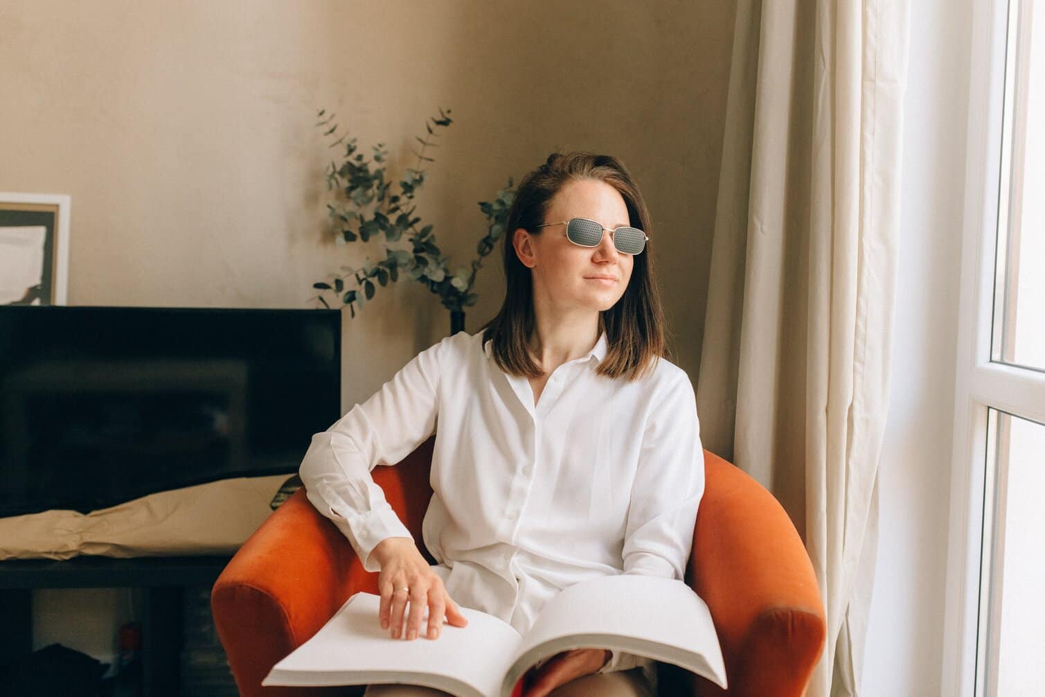 A lady with a white blouse and shoulder length dark hair, she is sat on a orange confutable chair. She is looking out of her window absorbing the sunlight. She has dark glasses on and has her left hand on her brail book. She is reading about the benefits of sight loss counselling.