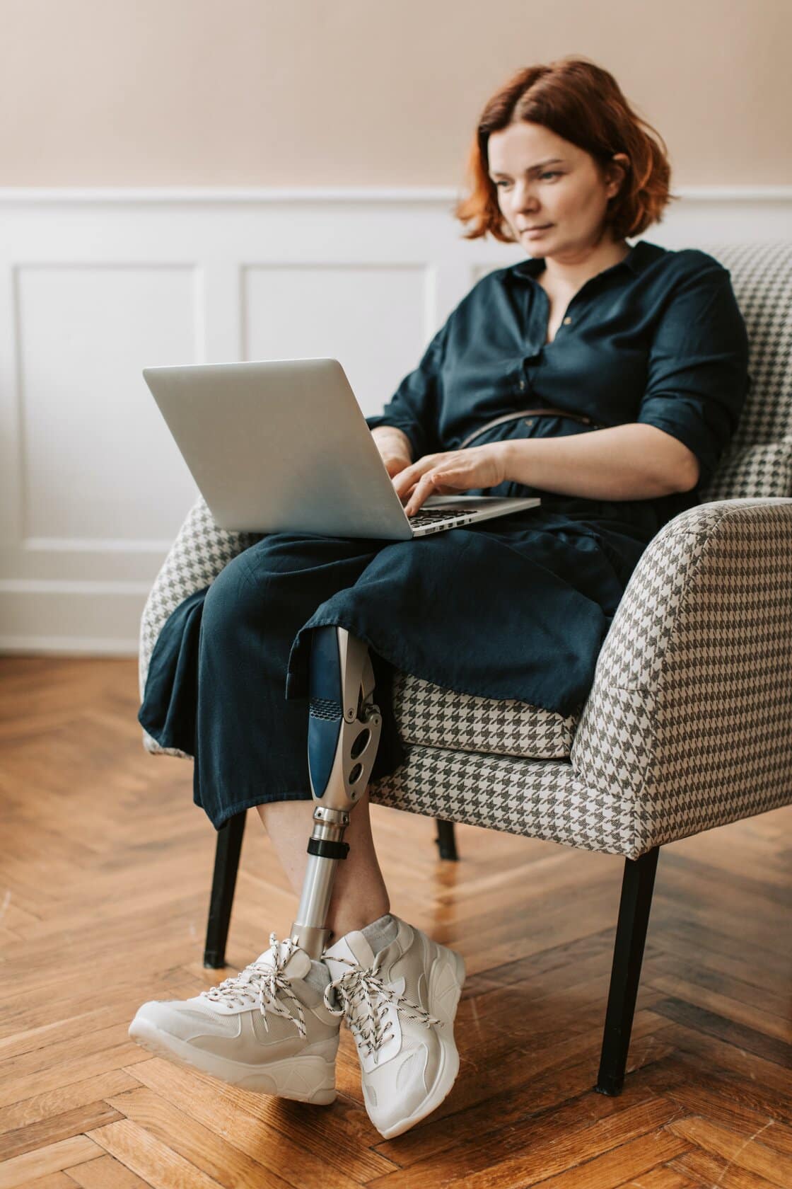 Lady sat in a chair, she has a lower limb amputation with prostecic leg. She is looking at her laptop, she is taking a loss of limb counselling session