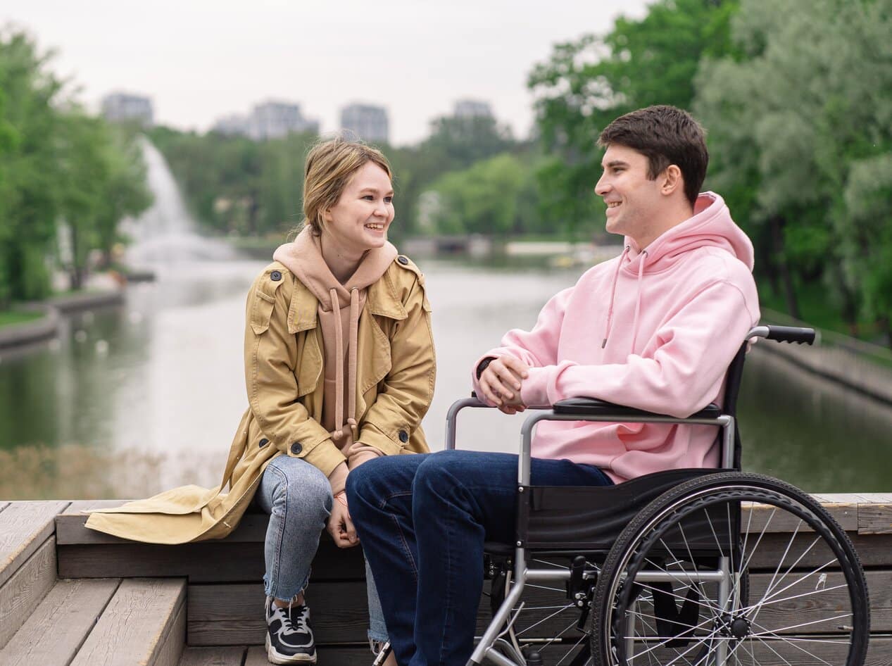 A man sat in his wheelchair in a park. He is sat with his partner and they are talking about spinal cord injury counselling