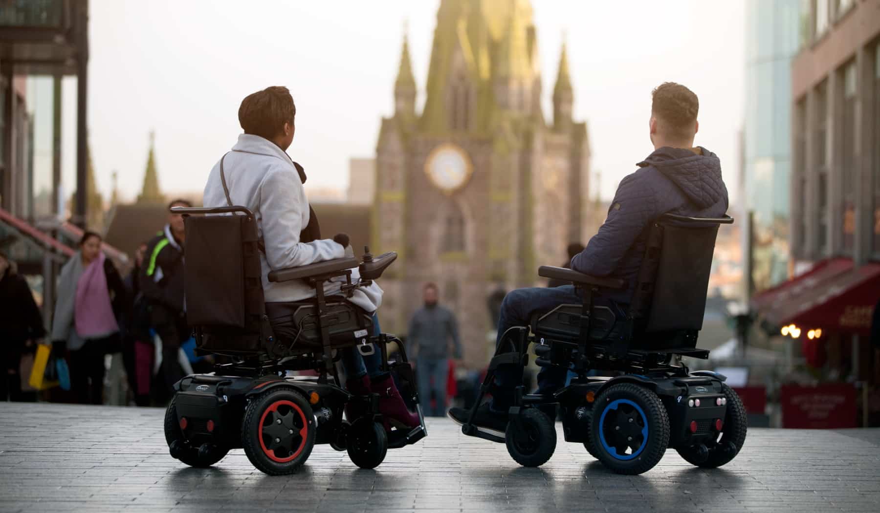 A couple sat in their wheelchairs on paving, they are looking at a church with many people walking towards them. They are feeling better after having spinal cord injury counselling