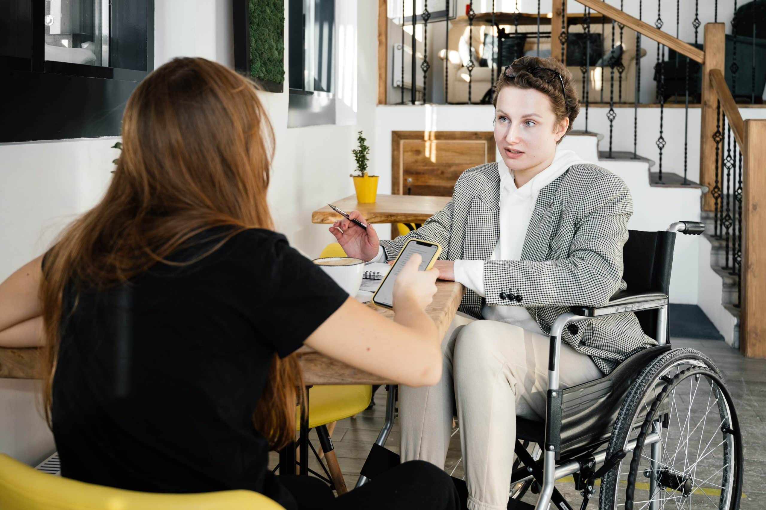 A lady sat in her wheelchair outside. She is sat next to her freind who is looking at the DisabilityPlus website, she is deciding if cerebral palsy counselling is good for her freind.