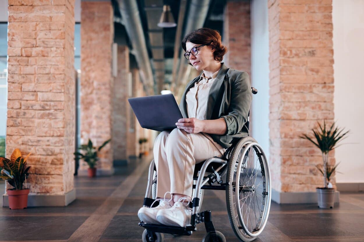 Lady sat at her table with large laptop open in front of her, she is saying high to a person who has started a disability counselling session.