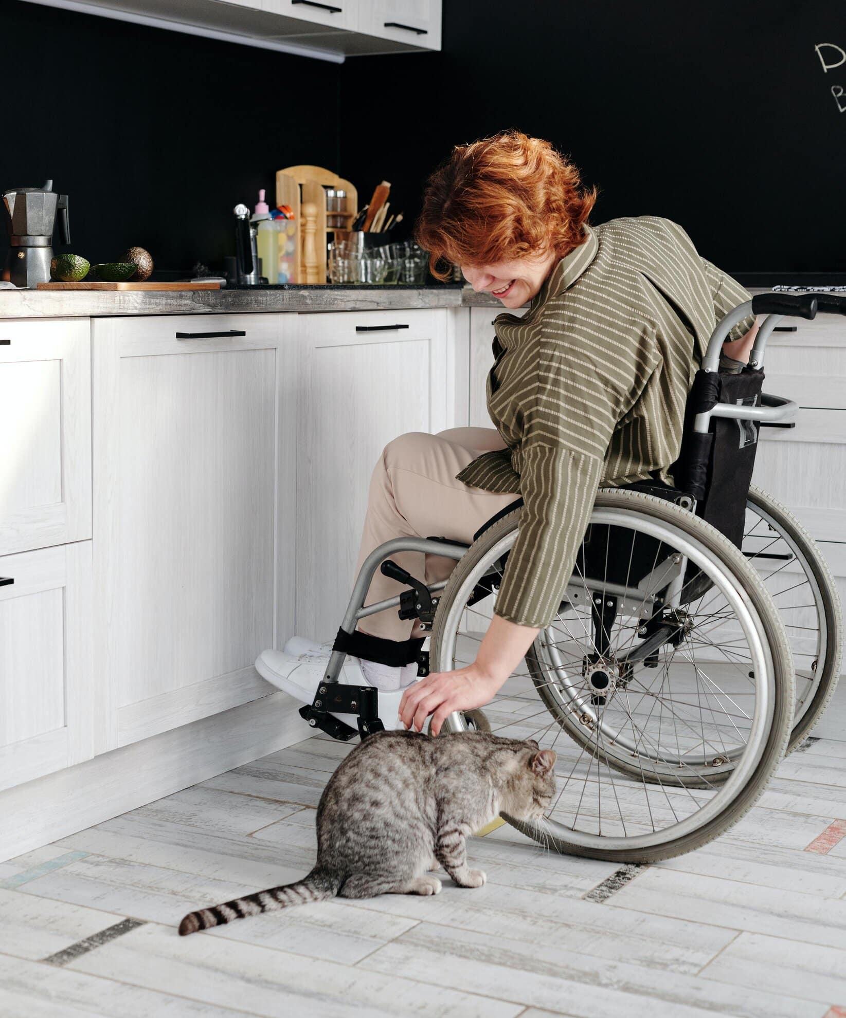 Lady sat in her wheelchair, she is in her kitchen stroking her cat.