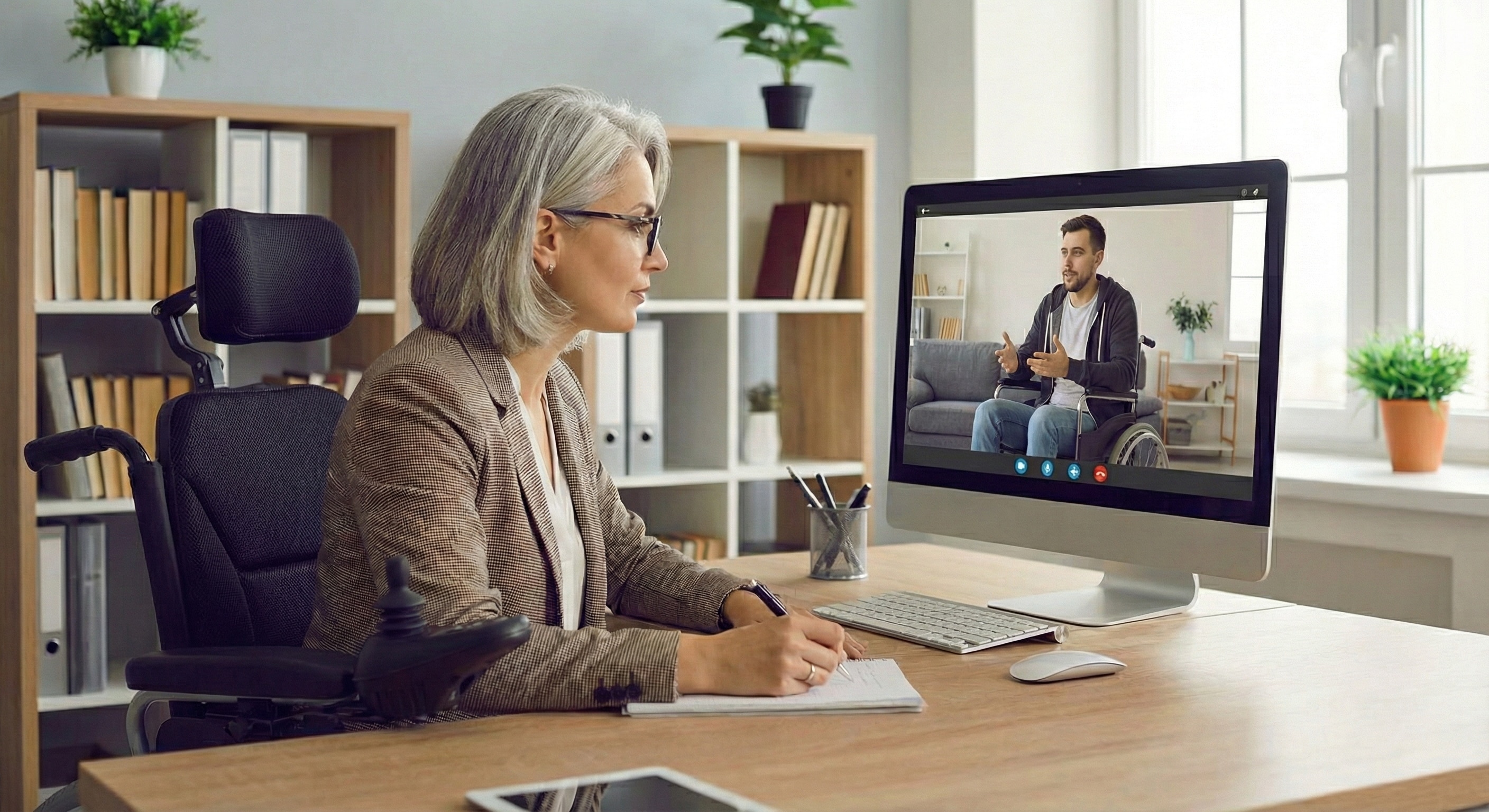 "A woman seated in a powered wheelchair at a well-lit desk, writing on a notepad as she is conducting a PIP Mental Health Assessment Report. She is looking at her large desktop monitor, which shows a video call with a man who is also using a wheelchair."